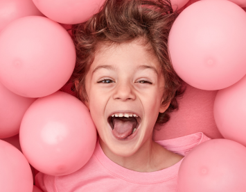 A joyful child with curly hair smiles widely while surrounded by pink balloons. Their playful expression conveys happiness in a vibrant, cheerful setting.