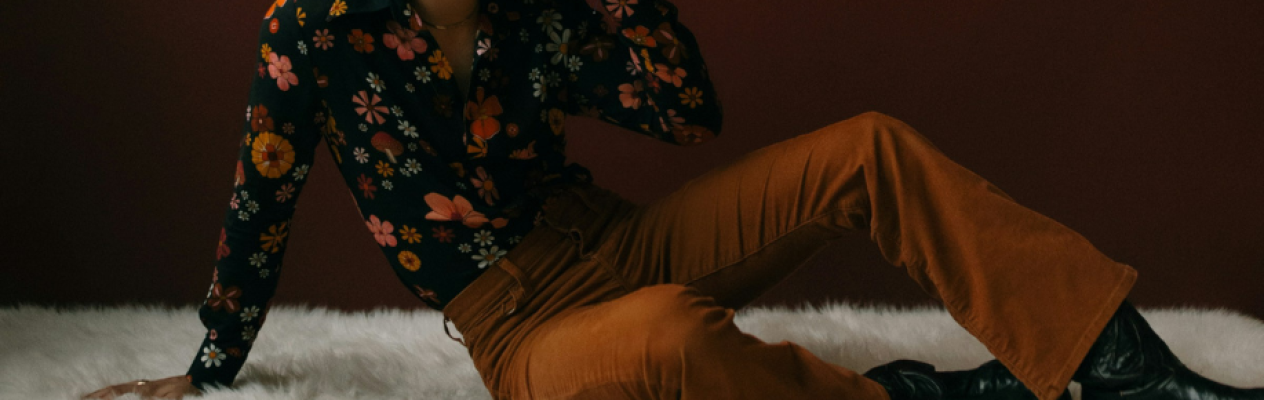 A person sits on a fluffy white surface, wearing a black floral-patterned shirt and brown flared pants. They have a stylish pose, looking away from the camera against a maroon background.