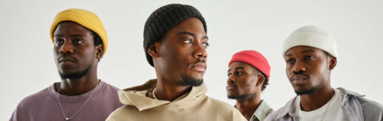 Four men wearing stylish beanies in various colors stand side by side against a plain white background. Each man has a distinct expression while dressed in casual, contemporary clothing.