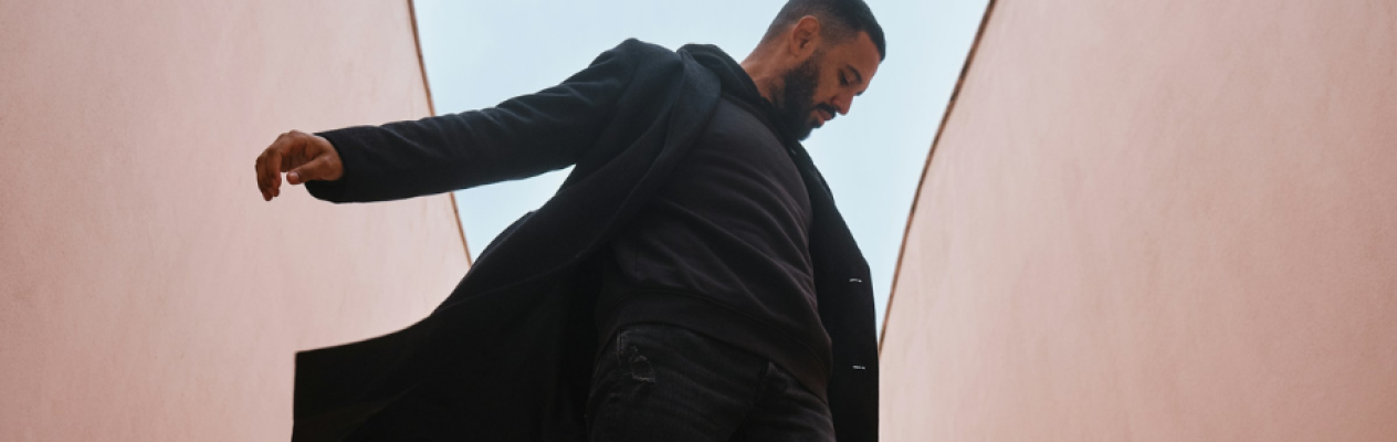 A man in a black coat and dark clothing stands in a narrow, pink-walled corridor, looking down with a thoughtful expression. The architecture creates a dramatic perspective emphasizing the height of the walls.