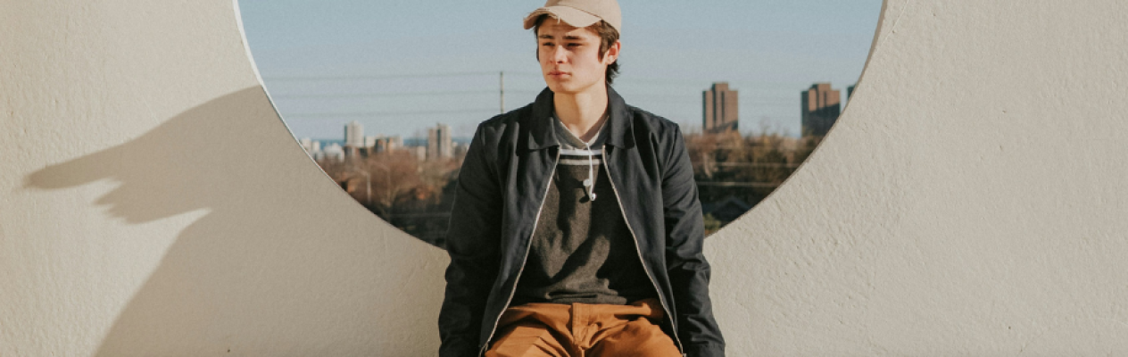 A young man sits casually on a ledge, framed by a circular opening in a white wall. He wears a black jacket, gray sweater, and tan pants, with a light baseball cap. The background features a city skyline under clear skies.