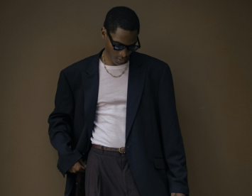A person wearing large sunglasses and a black blazer over a light-colored shirt stands with their head slightly bowed. They have a gold chain around their neck and are wearing dark trousers. The background is a muted brown color.