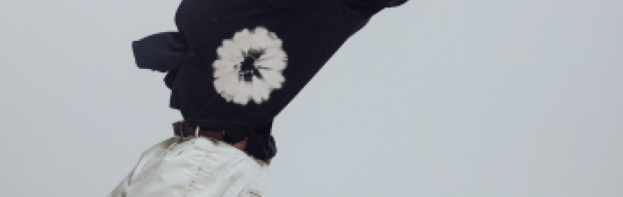 A person wearing a black tie-dye shirt and light-colored pants appears to be mid-dance, leaning back with one arm extended. They have glasses and a confident expression.