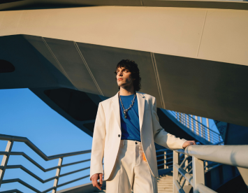 A person stands confidently on a staircase under a modern architectural structure. They wear a white suit over a blue t-shirt, accessorized with a statement necklace. The scene is illuminated by warm sunlight against a clear blue sky.