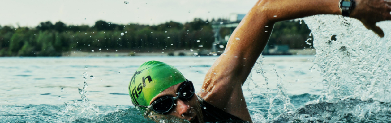 A swimmer in a green cap and sunglasses performs a freestyle stroke in a shimmering body of water, creating splashes around them. Lush trees are visible in the background under a cloudy sky.