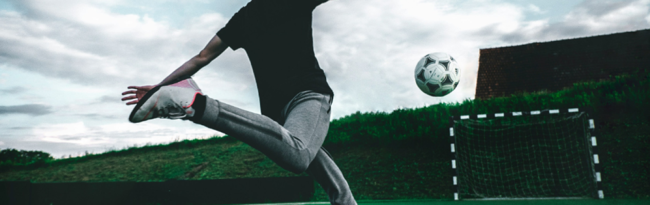 A young person in a black t-shirt and gray pants kicks a soccer ball on a grassy field, with a goalpost visible in the background under a cloudy sky.