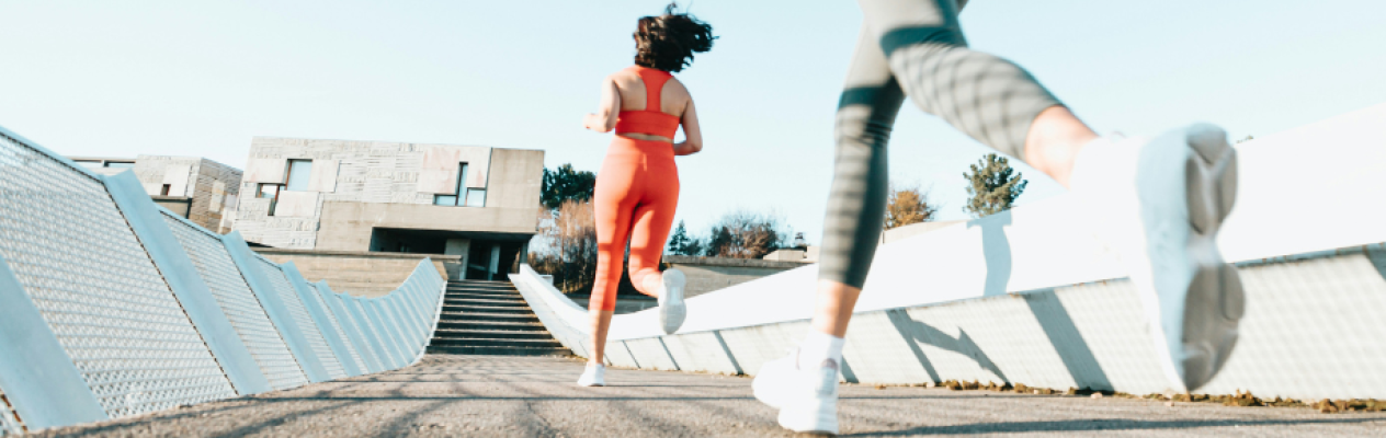 Two women jog on a path with a modern building in the background. One is wearing a red athletic outfit, while the other is in gray leggings and a matching top. Bright sunlight illuminates the scene, and the atmosphere is lively and active.