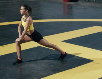 A woman in a yellow sports bra and black shorts performs a lunge on a black and yellow gym floor. Her hair is pulled back, and she wears black athletic shoes.