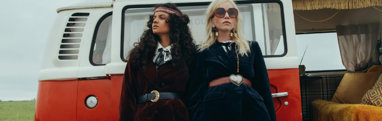 Two women stand confidently in front of a vintage red and white Volkswagen van. One has curly hair and wears a dark brown velvet outfit with a bow tie. The other has straight blonde hair, sunglasses, and a navy blue velvet suit with a heart-shaped belt buckle. The backdrop features green fields under a gray sky.