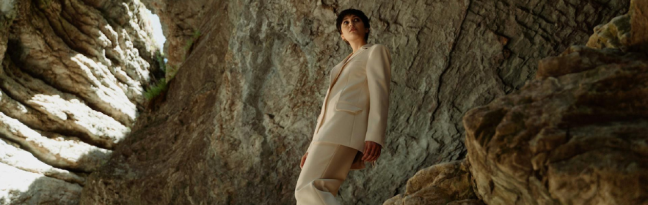 A person stands confidently on a rocky surface, dressed in a stylish beige suit. The backdrop features rugged, textured rocks with natural lighting highlighting the scene.