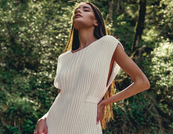 A woman poses confidently outdoors, wearing a stylish white dress with vertical pleats. Her long hair flows down, and she gazes upward with a serene expression, framed by lush greenery.