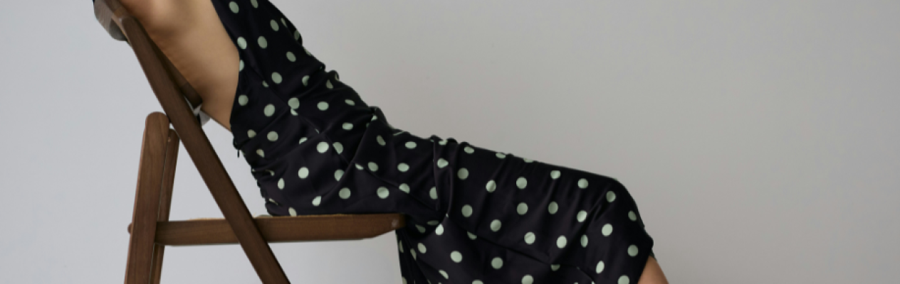 A person in a polka dot dress reclines on a wooden chair, creating a relaxed and stylish pose. The background is a neutral color, emphasizing the outfit.