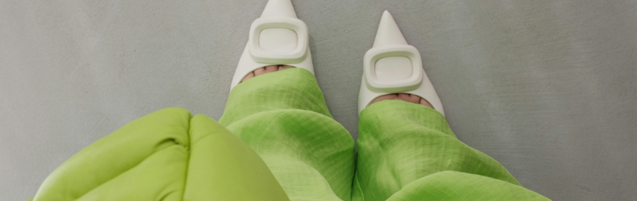 A pair of stylish white pointed-toe heels with square buckles is worn with flowing green trousers. The view is from above, showcasing the shoes and the fabric's texture.
