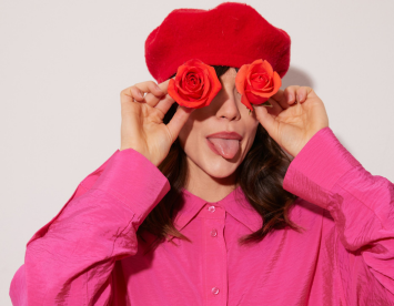 A person playfully poses with a bright red beret and holds orange roses in front of their eyes while sticking out their tongue. They are wearing a vibrant pink shirt.