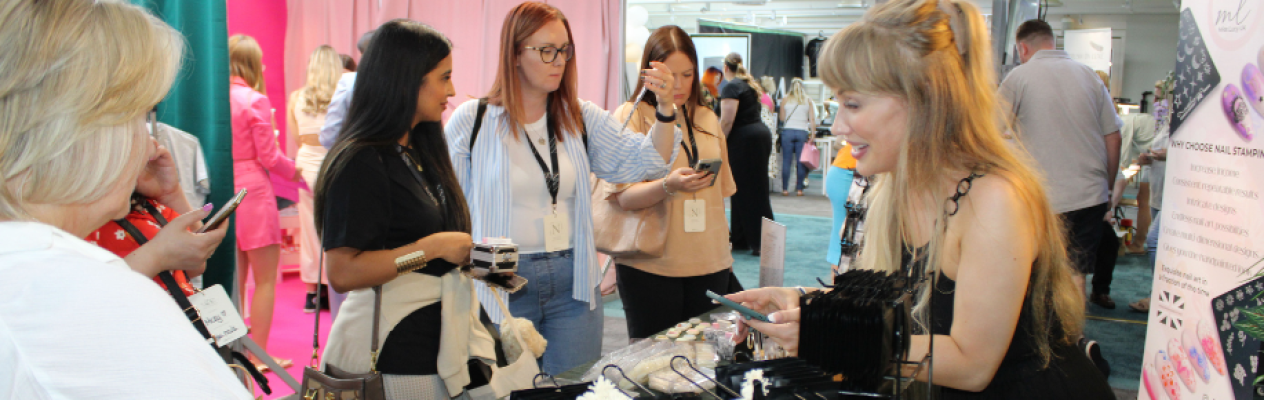 A group of women gathers around a vendor at a trade show. One woman at the booth demonstrates products, while others engage with their phones, expressing interest in the displayed items. Colorful backdrops create a vibrant atmosphere.