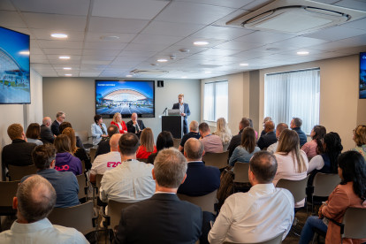 A speaker stands behind a podium in a conference room, addressing an audience seated in rows. The room features a large screen displaying a presentation, with several panelists seated to the left. Natural light streams in through windows.