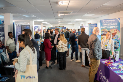A busy event space filled with attendees networking and discussing at various booths. Tables display promotional materials, with individuals engaging in conversations throughout the area.