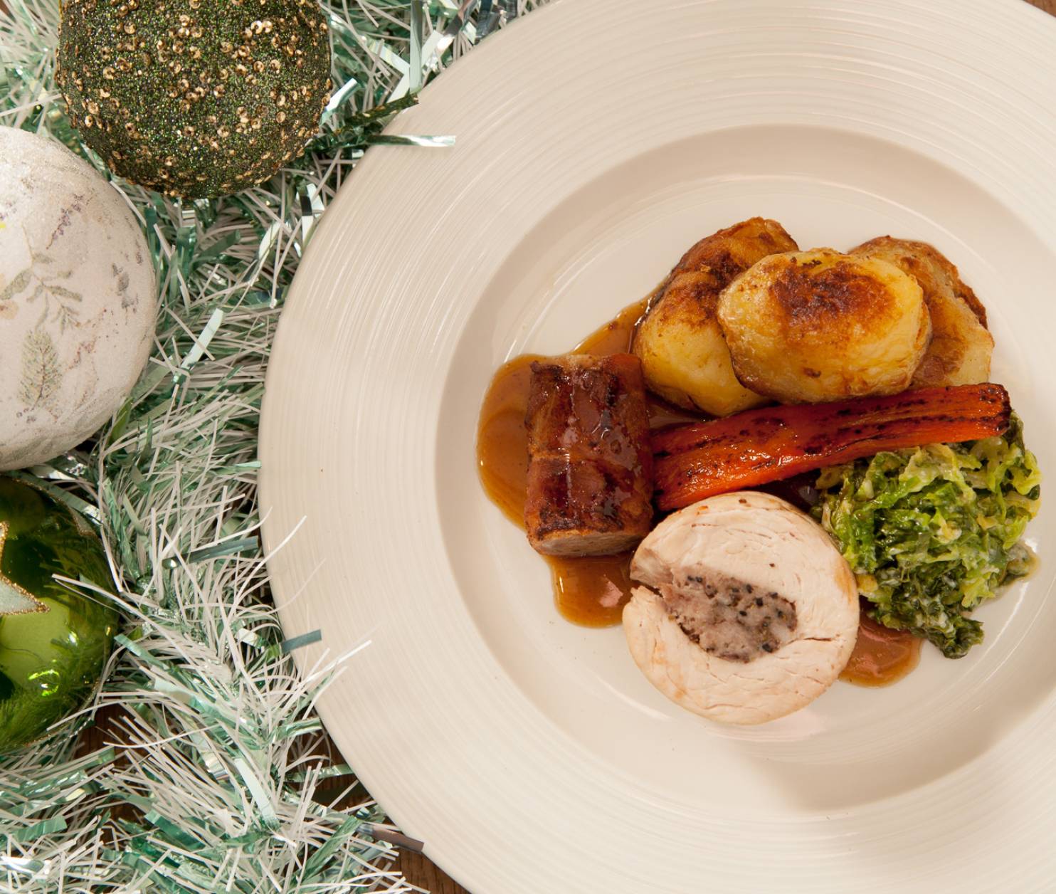 A festive plate of holiday dinner featuring sliced turkey roll with stuffing, roast carrots, golden roast potatoes, and green vegetables, drizzled with gravy. Surrounding the plate are decorative Christmas ornaments in green and white, along with shiny tinsel.