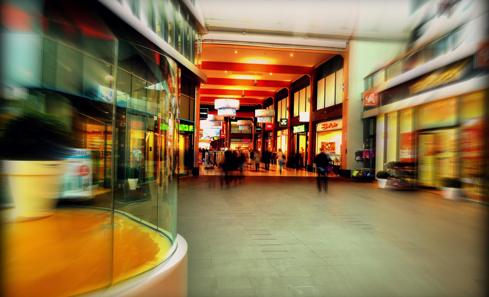 A bustling shopping mall interior with blurred figures walking through. Brightly lit storefronts line the corridor, showcasing colorful signs and promotions. The atmosphere is lively, with modern architecture and decorative elements.