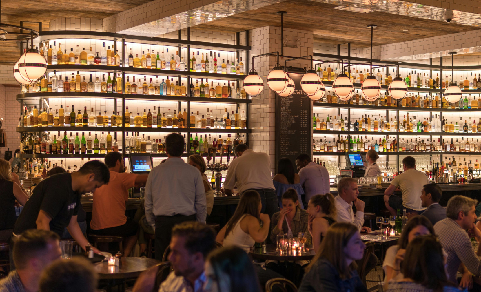 A lively bar scene with patrons enjoying drinks. Shelves filled with various bottles of liquor are well-lit in the background, and people are seated at tables, some engaging in conversation, creating a vibrant atmosphere.