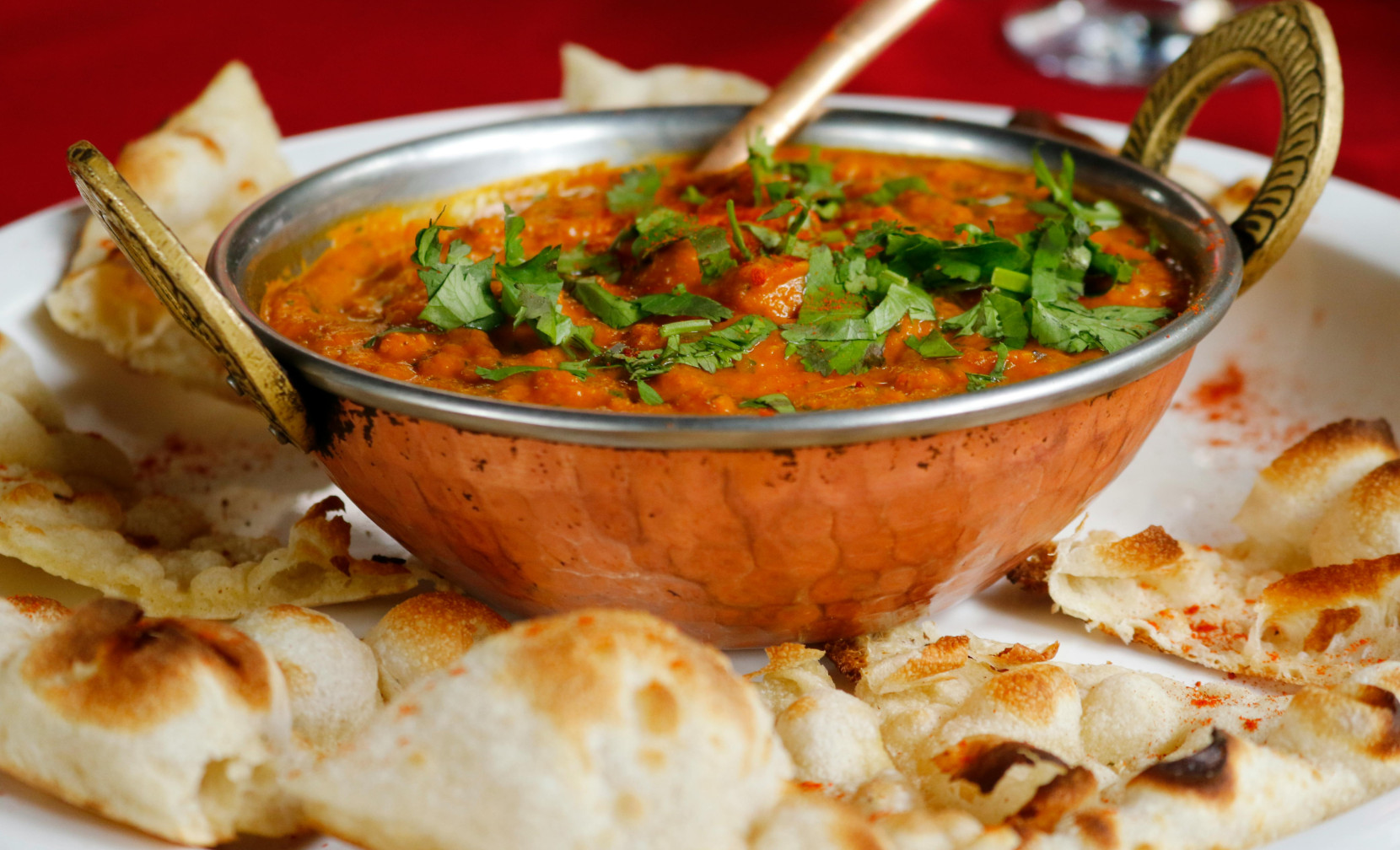 A copper bowl filled with a rich, orange curry topped with fresh cilantro, surrounded by pieces of naan bread on a red tablecloth.