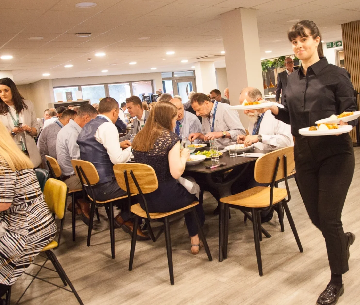 A waitress in a black uniform carries two plates of food while a dining area filled with people enjoying a meal is visible in the background. Some guests are engaged in conversation, seated at tables with modern chairs.