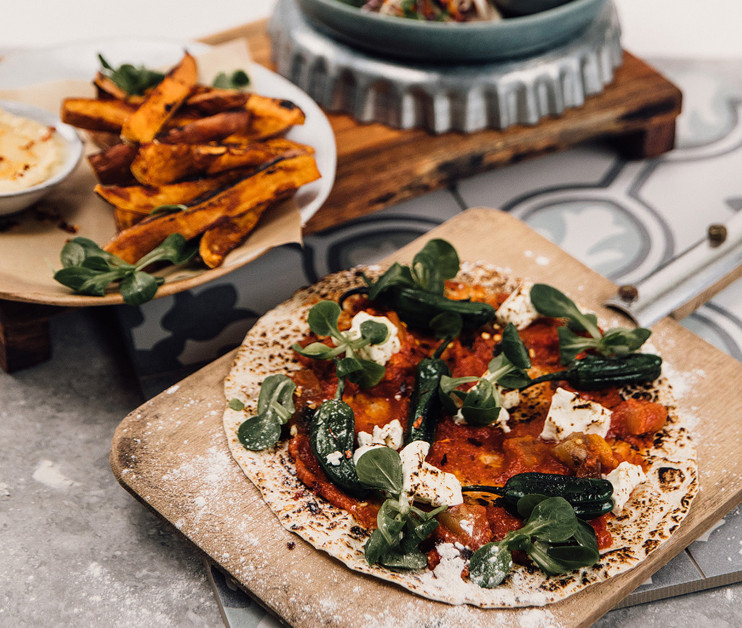 A wooden board holds a freshly made flatbread topped with tomato sauce, feta cheese, and fresh greens, alongside a serving of sweet potato fries and a small bowl of dipping sauce.