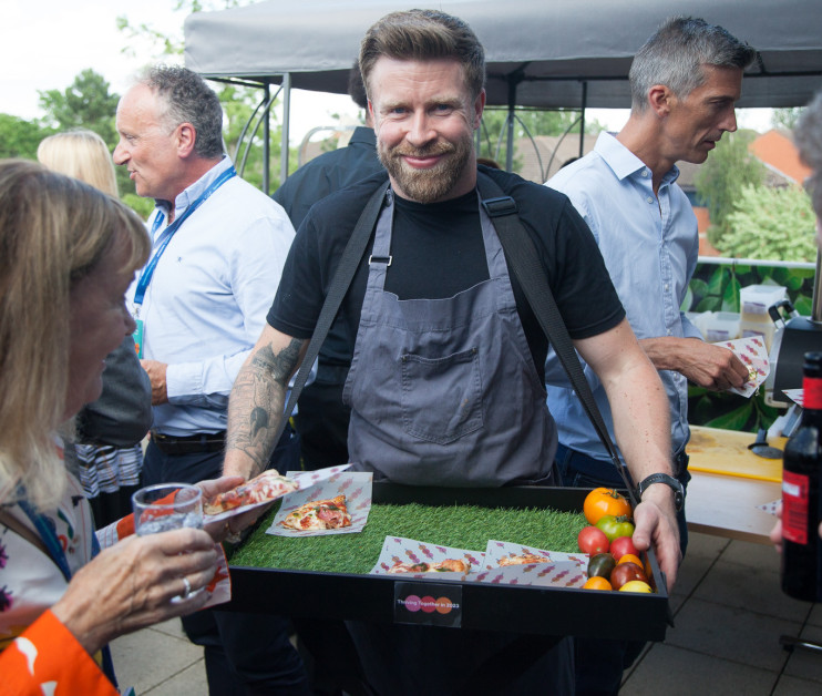 A man with a beard and tattoos wears an apron and smiles while holding a tray of food, including pizza slices and fruits, at an outdoor event with attendees mingling around him.
