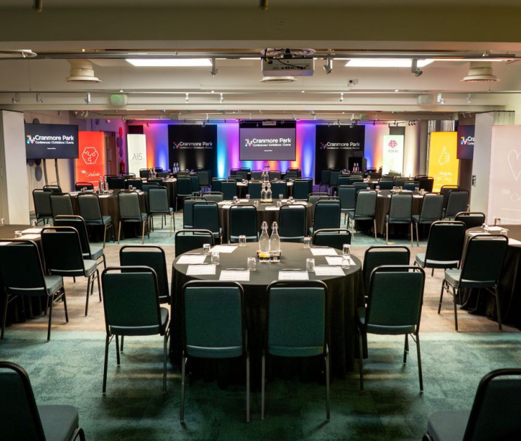 A conference room set up with multiple rows of tables and chairs, featuring branding banners along the walls. Two screens are displayed at the front, and the lighting creates a vibrant atmosphere.