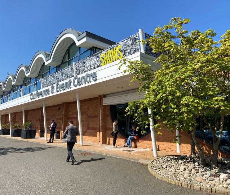 A modern conference and event center showcasing a distinctive curved roof design. People are entering and exiting through the main entrance, which features large glass doors. Lush greenery and decorative stones are positioned around the building's exterior.
