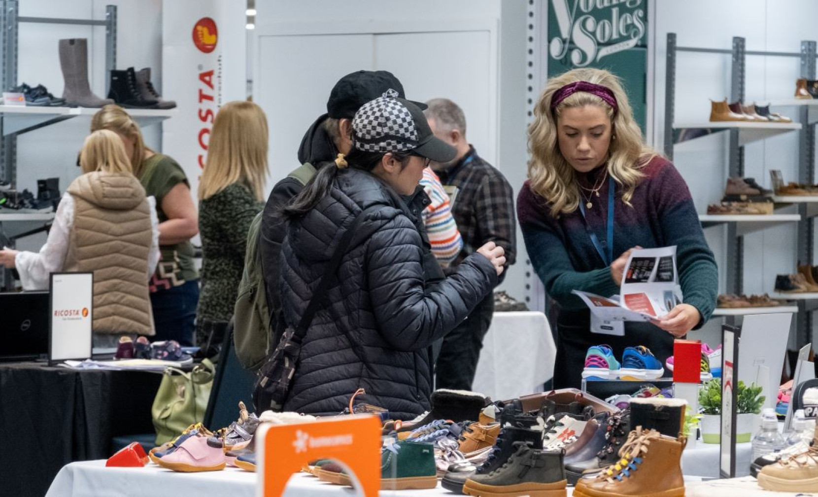 A group of people shopping at a footwear exhibition with various shoe styles on display. One woman is examining a brochure while another browses. In the background, others engage with the booths and products.