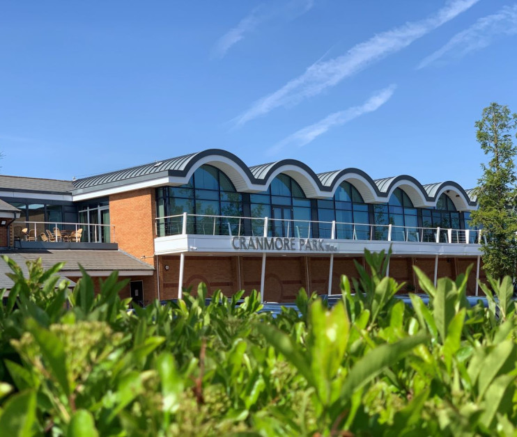 Cranmore Park building with a curved roof design, featuring large windows and a balcony. Clear blue sky and greenery in the foreground.