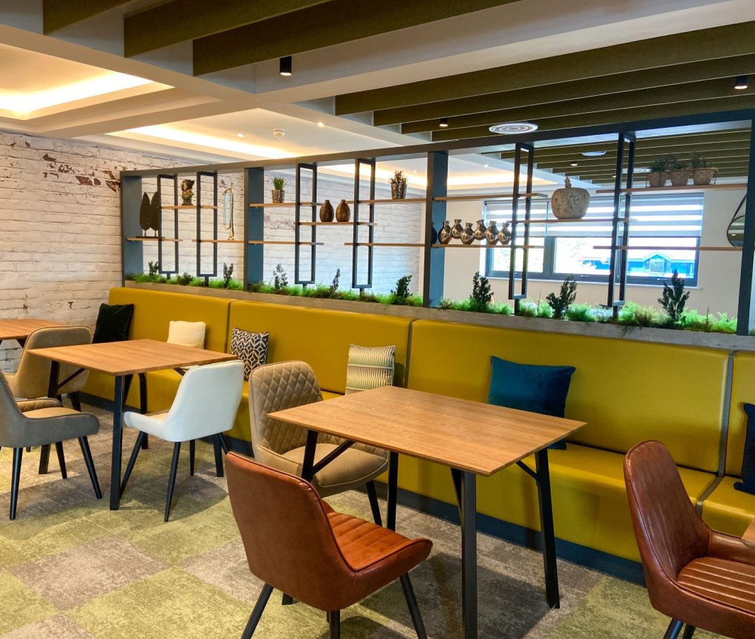 A modern cafe interior featuring wooden tables and a variety of seating options, including upholstered chairs. Greenery accents the space, with shelves displaying decorative items in the background. Soft lighting enhances the inviting atmosphere.