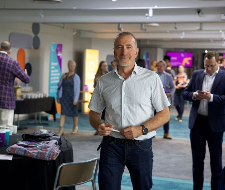 A man in a light blue shirt stands confidently in a modern event space, smiling at the camera. In the background, attendees engage in conversation and coffee service is visible. Bright banners decorate the room, adding to the lively atmosphere.