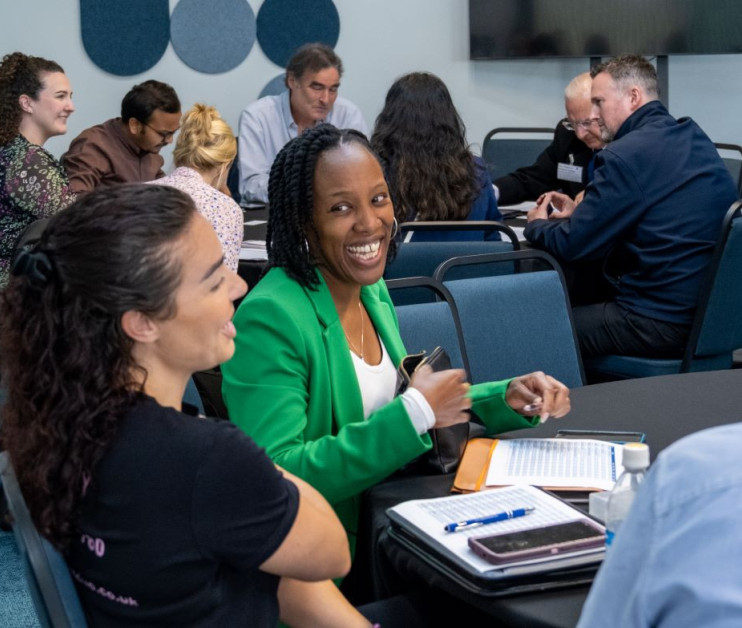 A diverse group of people engaged in discussion around a conference table. In the foreground, a woman in a green jacket smiles at the camera while seated next to another woman. Other participants are seen in the background, participating in a collaborative meeting.