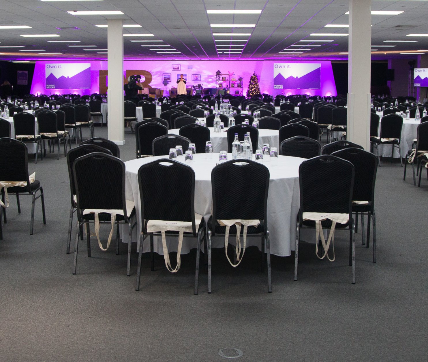 A spacious conference hall set up with multiple round tables, each covered with white tablecloths and equipped with water bottles and glasses. The room features a stage at the front with purple lighting and screens on either side.