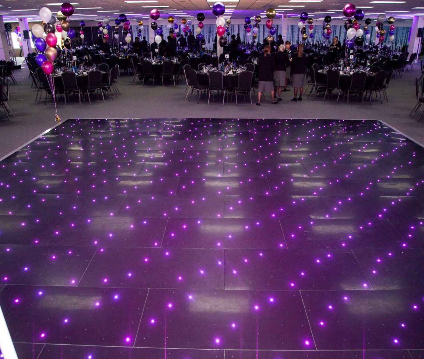 A spacious event hall is decorated with colorful balloons and elegant tables set for a celebration. The dance floor features a sparkling pattern of purple lights that adds a festive ambiance.