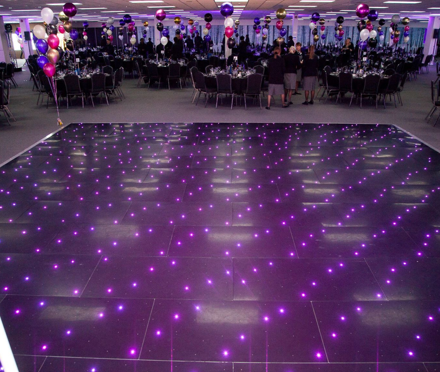 A spacious event hall is decorated with colorful balloons and elegant tables set for a celebration. The dance floor features a sparkling pattern of purple lights that adds a festive ambiance.