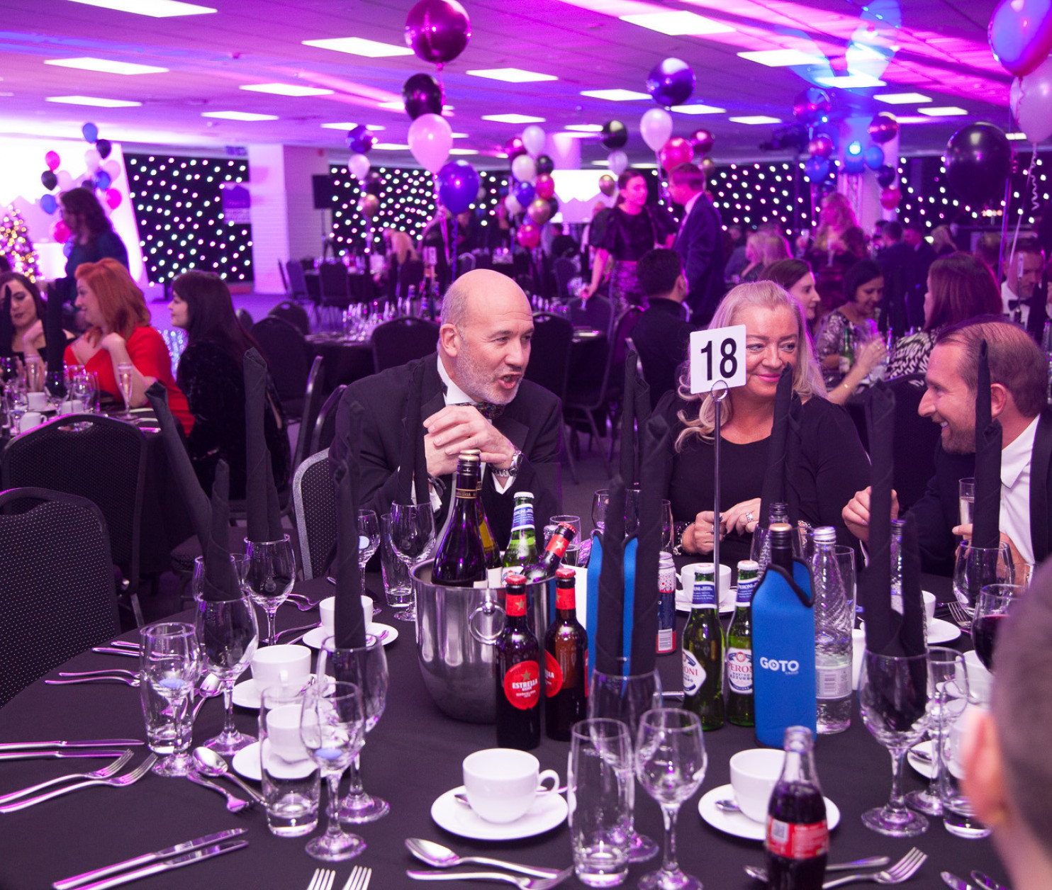 A lively banquet scene with guests seated at elegantly set tables. Colorful balloons and lights create a festive atmosphere, while people engage in conversation. Various drinks are displayed on the tables, adding to the celebratory vibe.