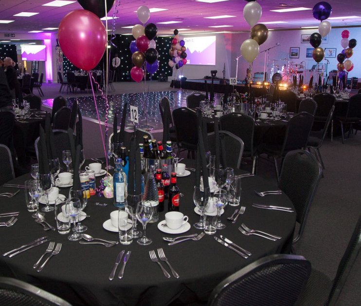 A banquet setup featuring a round table with black tablecloth, glassware, cutlery, and a variety of drinks. The room is decorated with balloons in pink, gold, and black, and a stage area with musical instruments can be seen in the background.