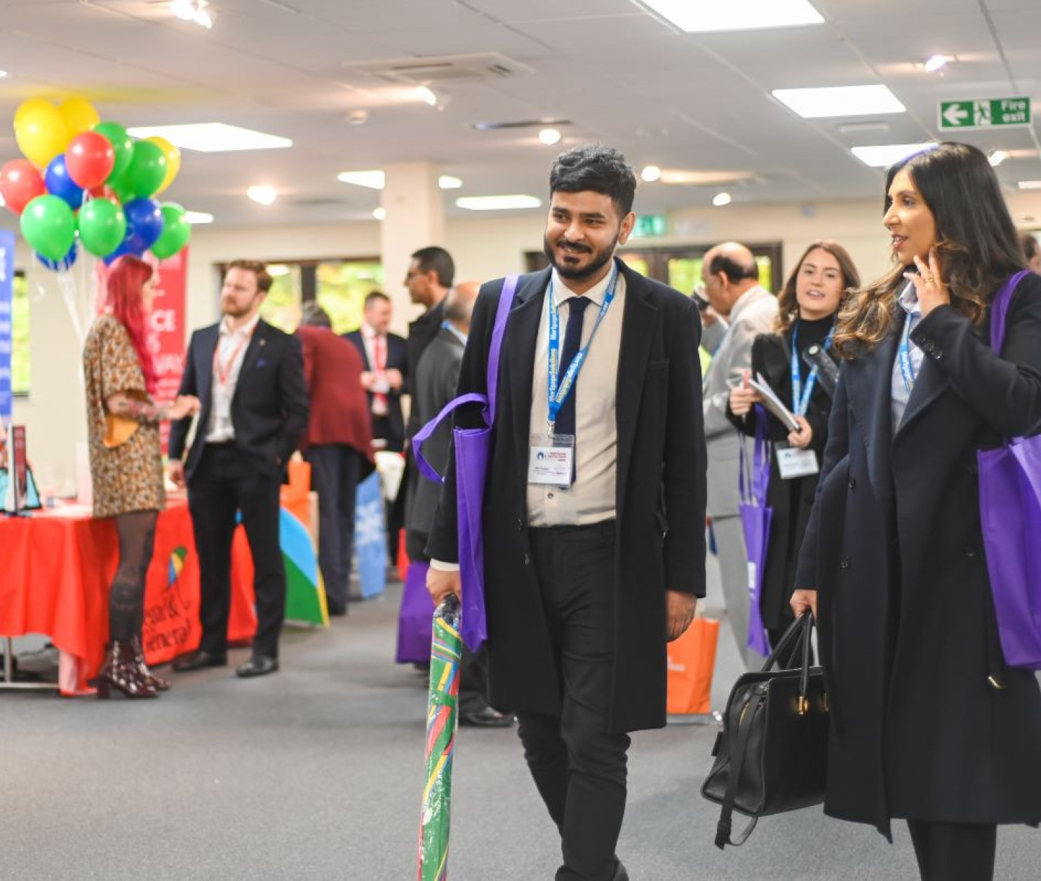 A group of professionals mingles at a networking event, with individuals carrying colorful bags. Balloons are in the background, and various exhibitor tables are set up around the venue.
