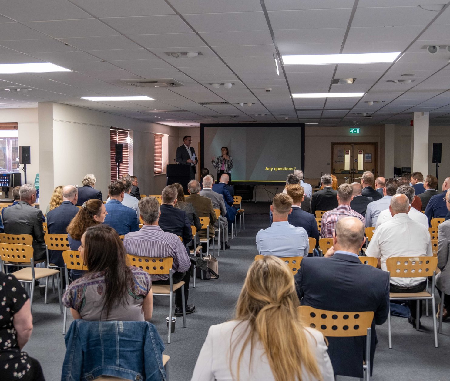 A crowded conference room with attendees seated, facing a presentation screen. Two speakers stand at the front, engaging the audience. Various people in business attire are positioned throughout the room, with a mix of men and women visible.