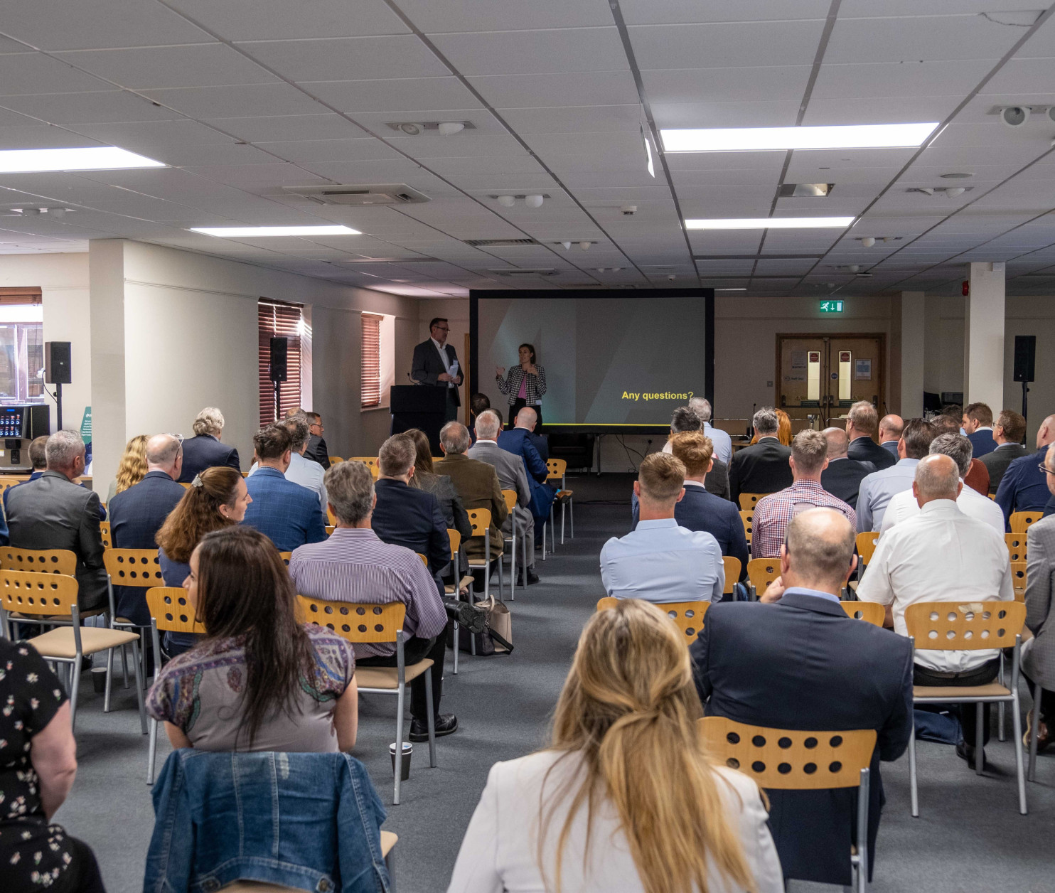 A crowded conference room with attendees seated, facing a presentation screen. Two speakers stand at the front, engaging the audience. Various people in business attire are positioned throughout the room, with a mix of men and women visible.