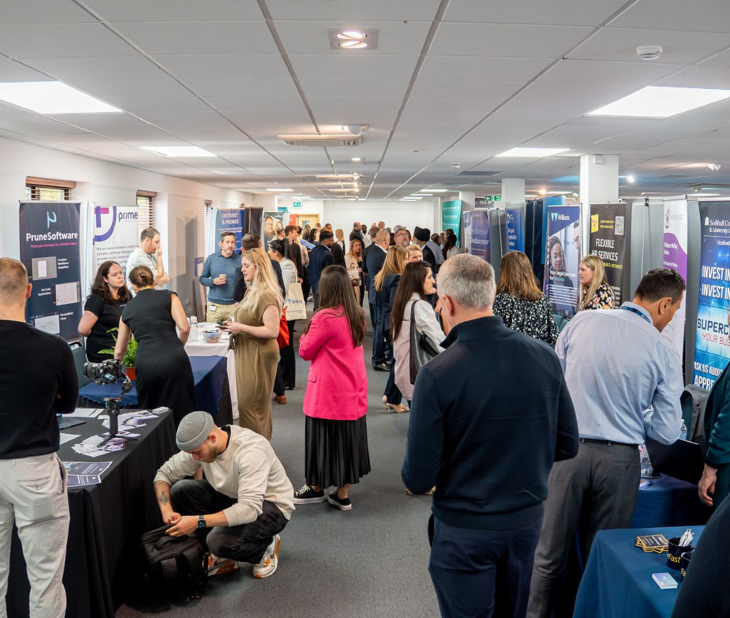 A bustling conference hall filled with people engaging at various booths. Attendees are networking and exploring displays about different programs and initiatives. Banners and signs promote services and skills training.
