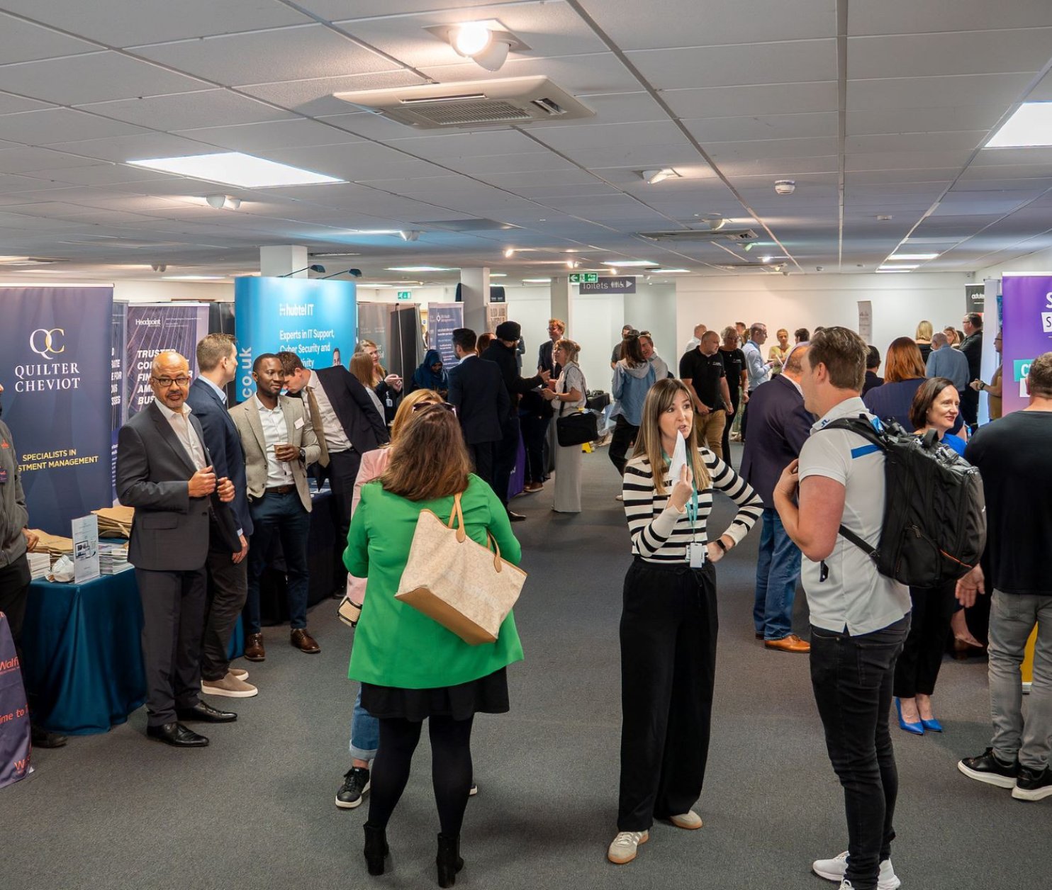 A bustling event space filled with people networking and engaging at various booths. Attendees are dressed in professional attire, with banners and tables showcasing different businesses and services.