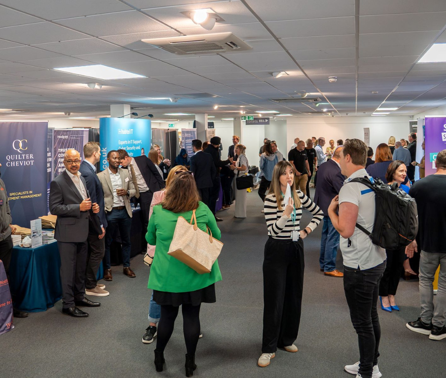 A bustling event space filled with people networking and engaging at various booths. Attendees are dressed in professional attire, with banners and tables showcasing different businesses and services.