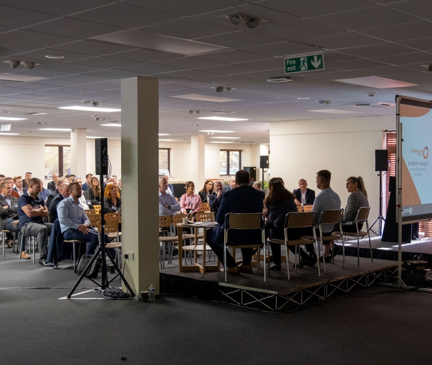 A panel discussion taking place in a well-lit conference room. Several participants are seated on stage, facing an audience. The audience is partially visible, engaged and attentive. A large screen displays text related to the event.