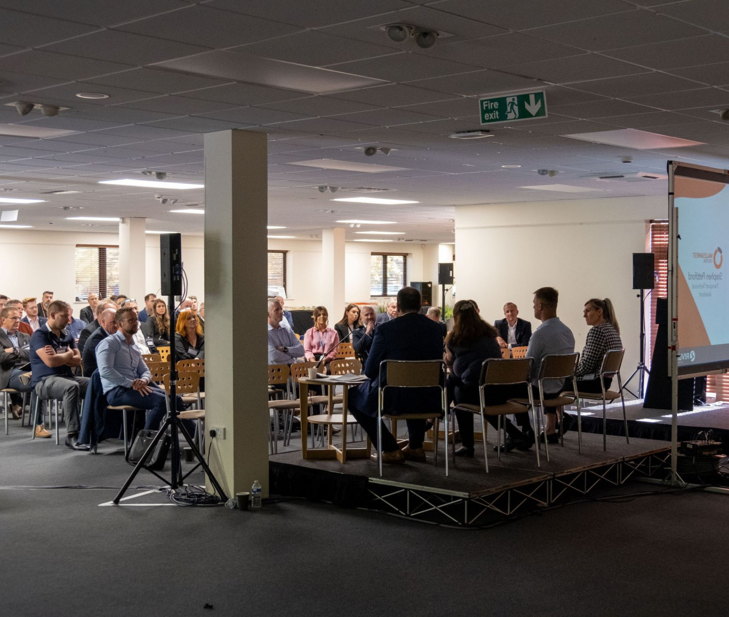 A panel discussion taking place in a well-lit conference room. Several participants are seated on stage, facing an audience. The audience is partially visible, engaged and attentive. A large screen displays text related to the event.