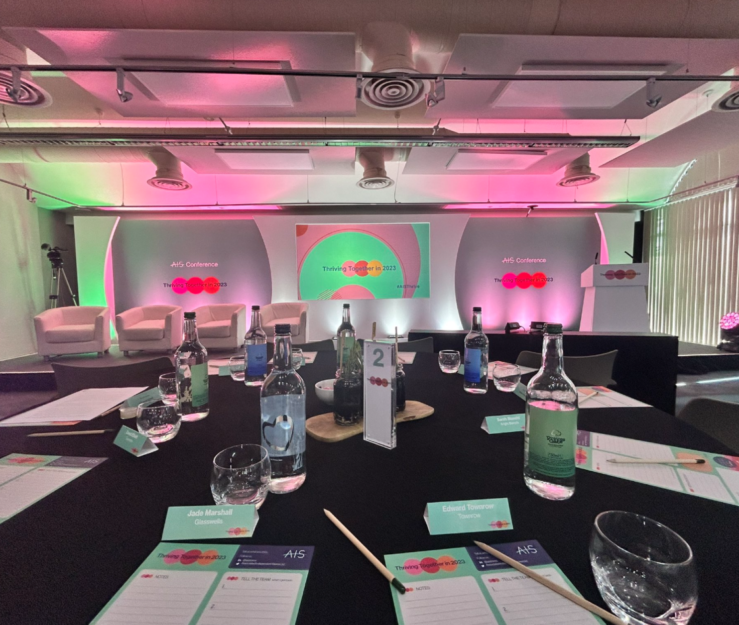 A conference room setup featuring a round table with notepads and pens. In the background, there is a colorful stage with lighting, equipped with a large screen and chairs arranged for a presentation.