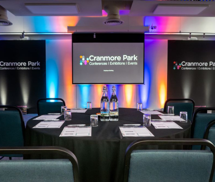 A conference setup with a round table covered in a black cloth, featuring bottles of water and glasses. Colorful ambient lighting enhances the space, with banners displaying 